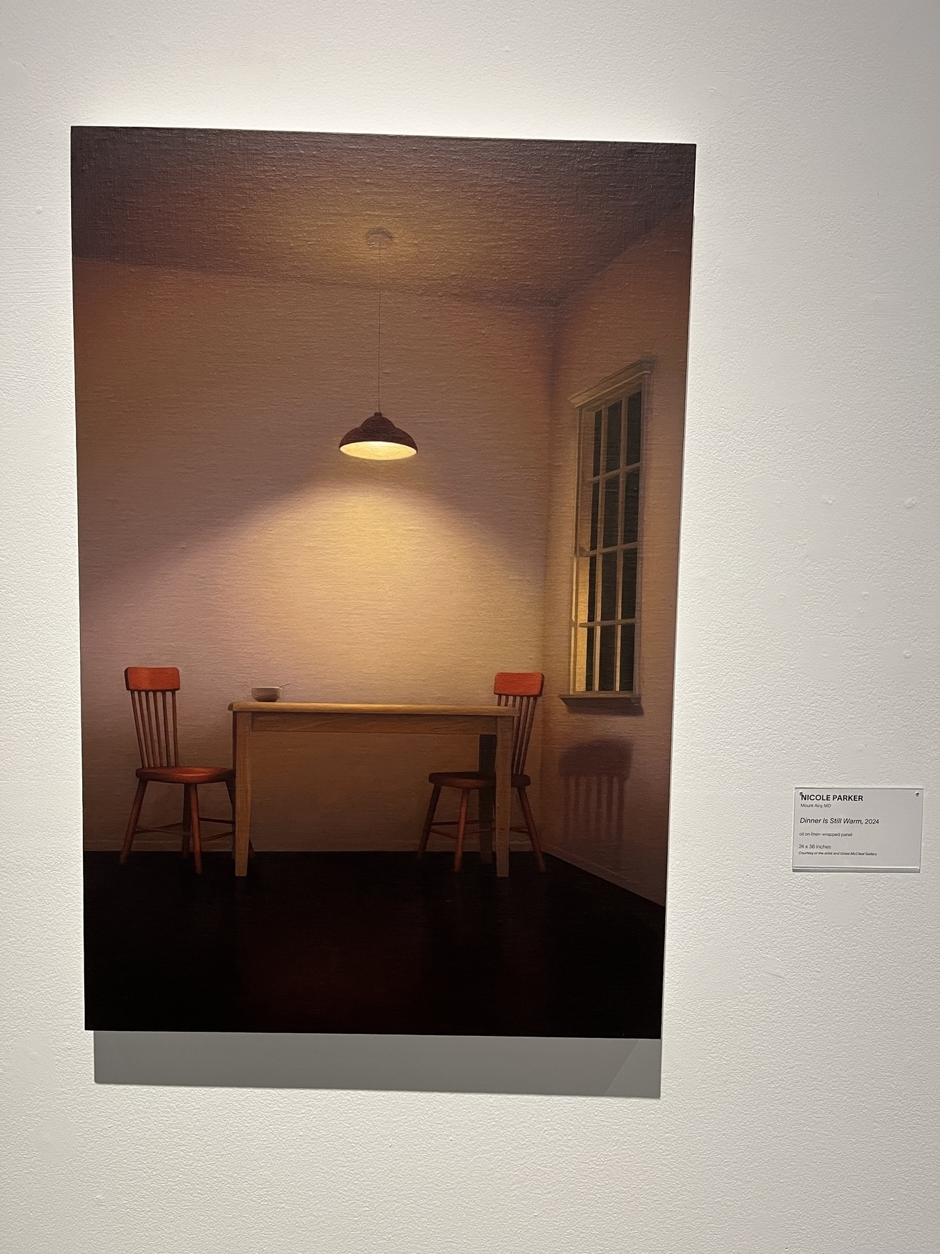 A wooden table with two chairs sits under a lit pendant light in a dimly lit room; a window casts a shadow. Text reads: “Nicole Parker, Dinner is Still Warm, 2021, oil on linen mounted panel.