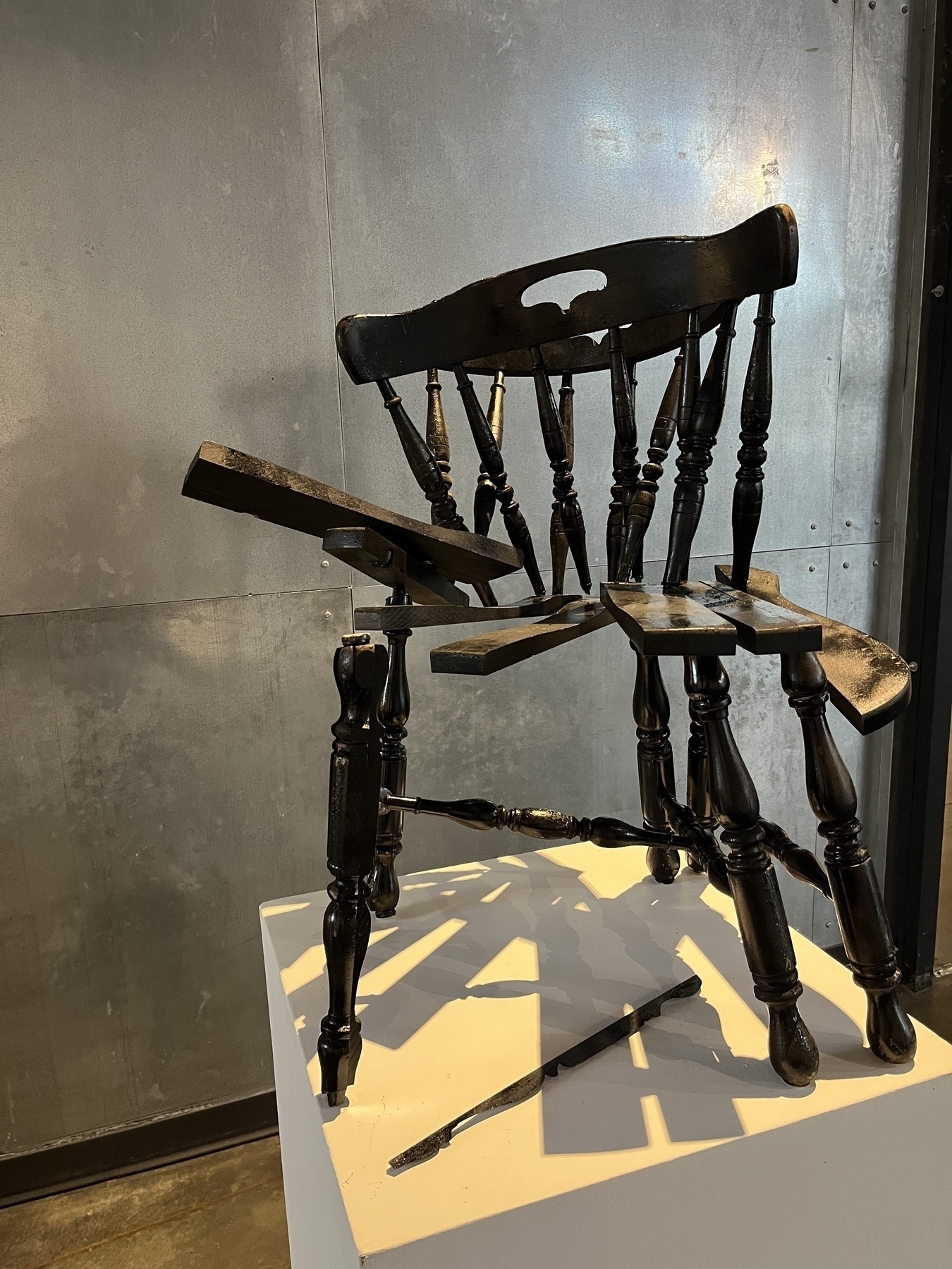 A broken wooden chair is precariously positioned on a white pedestal, with its legs akimbo and pieces scattered. It is set against a metallic, industrial-looking wall.