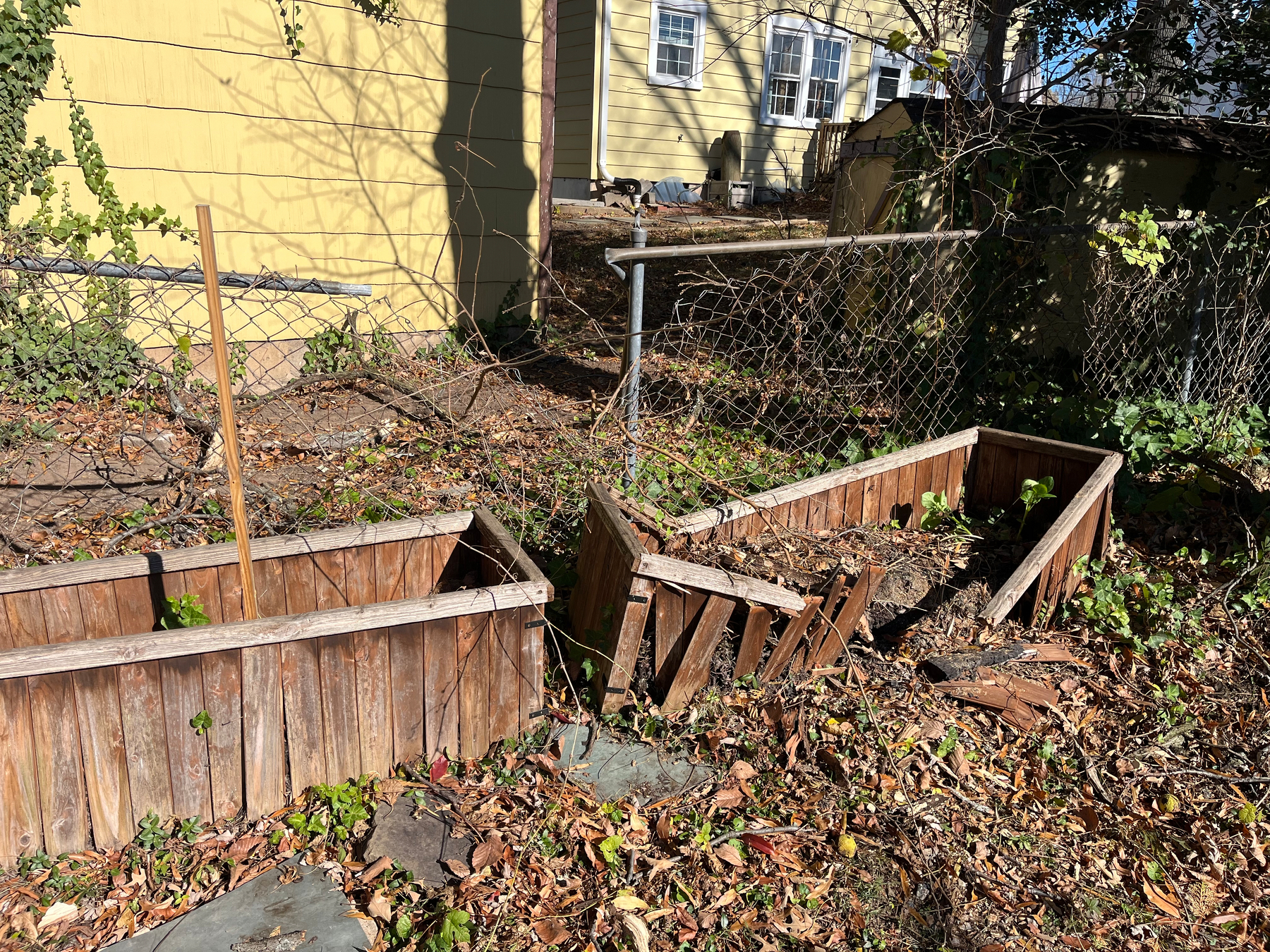 Two wooden planter boxes, one damaged, lie on a leaf-covered ground near a damaged chain-link fence. 
