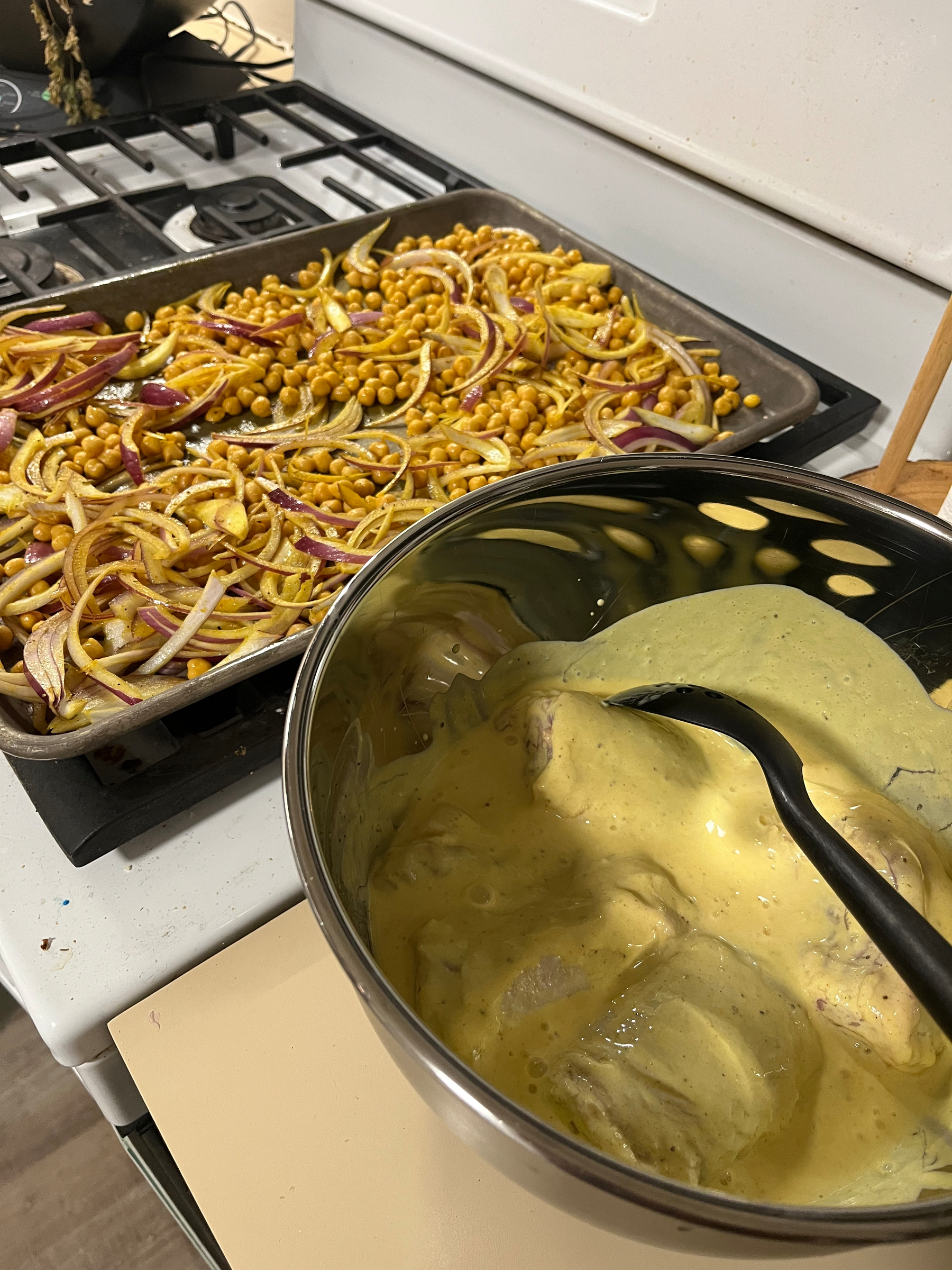 Chickpeas and red onions are spread on a baking tray atop a stove, while chicken pieces marinate in a yellow sauce in a metal bowl with a black spoon.