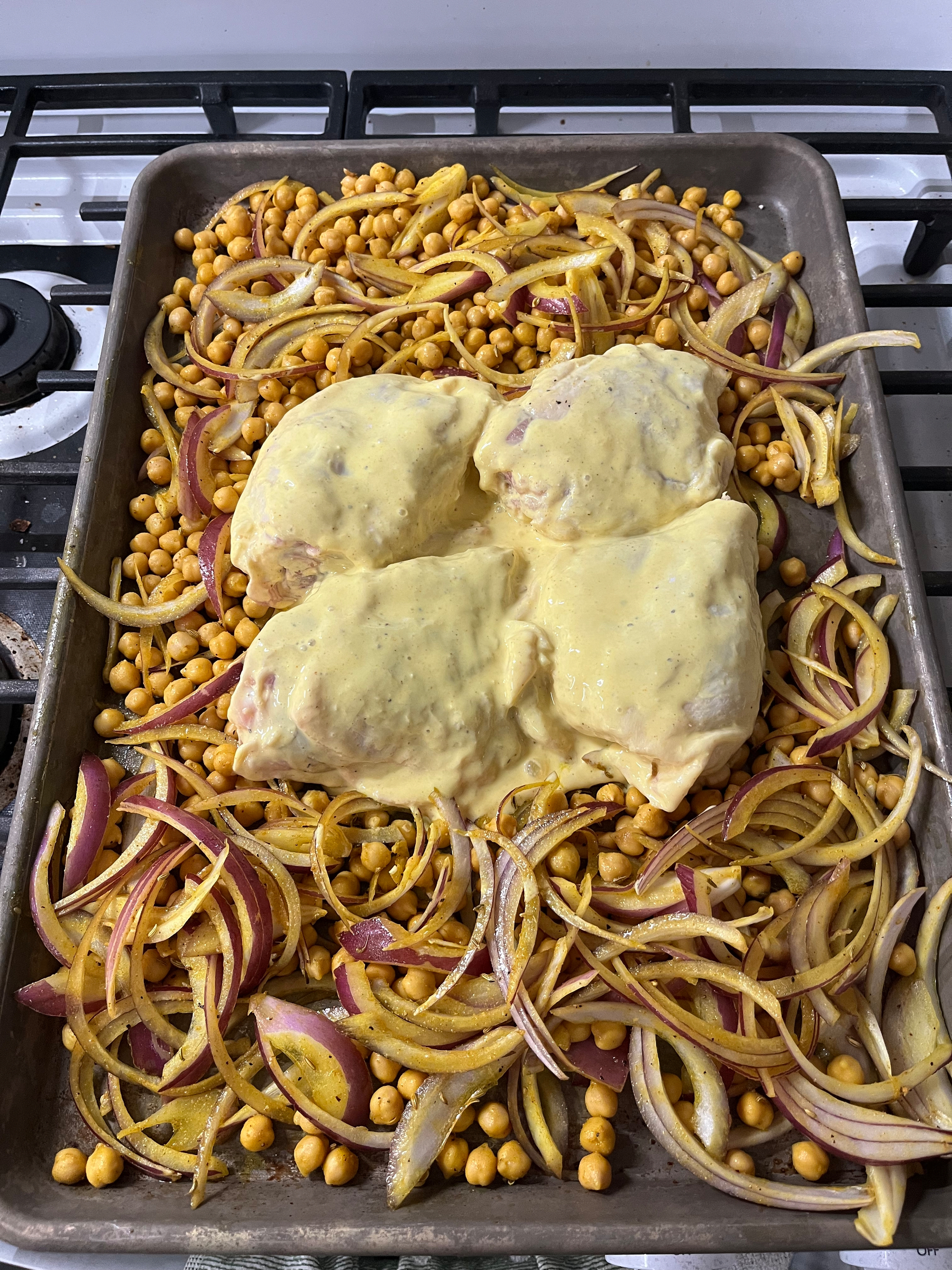 Chicken pieces coated in yellow sauce bake on a sheet pan with chickpeas and sliced red onions, resting on a stove.