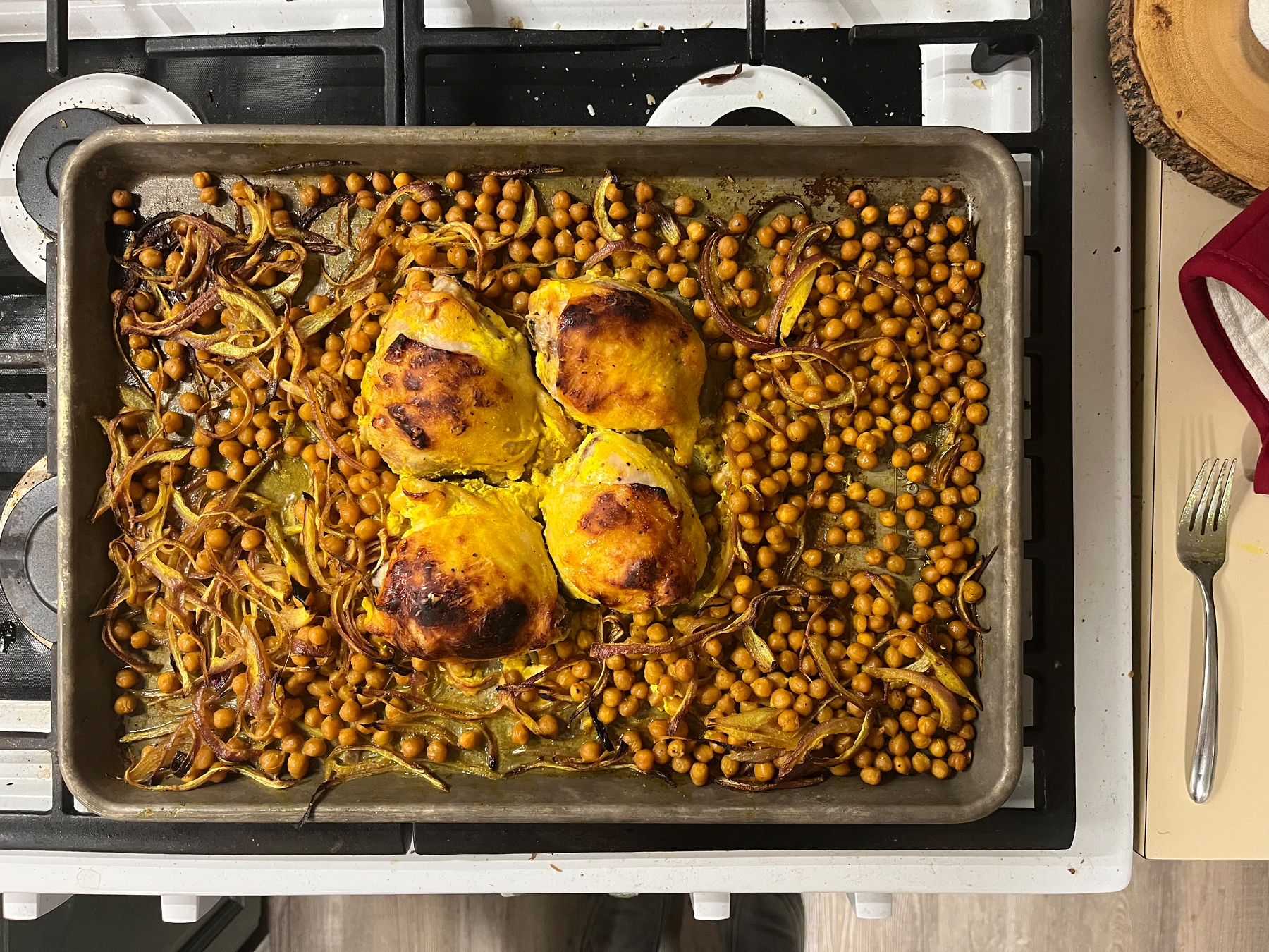 Roasted chicken thighs, surrounded by browned chickpeas and caramelized onions, rest in a baking tray. The tray sits on a stove with a wooden cutting board and fork nearby.