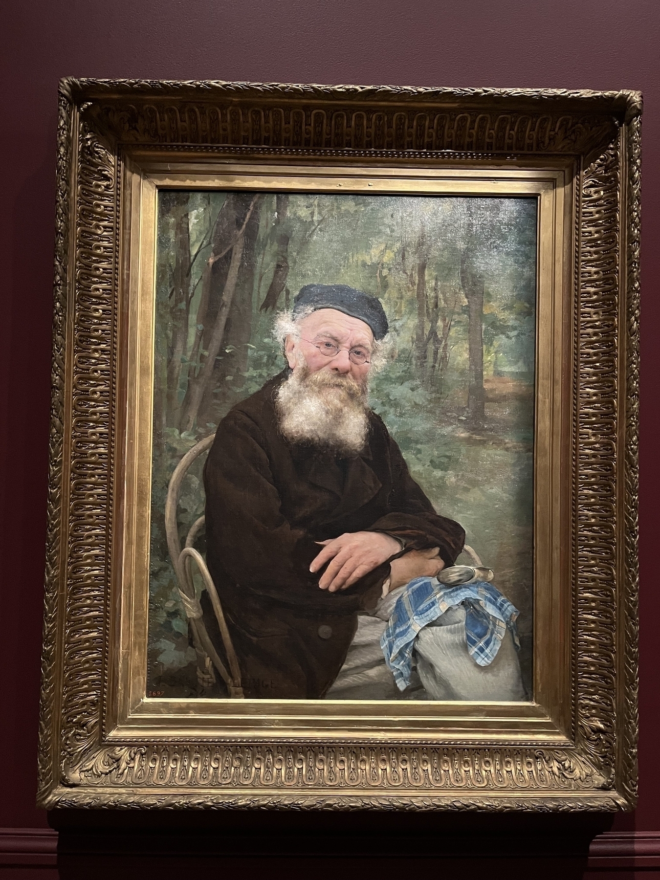 A seated man with a white beard rests hands on a knee, wearing glasses and a dark hat, set against a forest background inside an ornate gold frame.