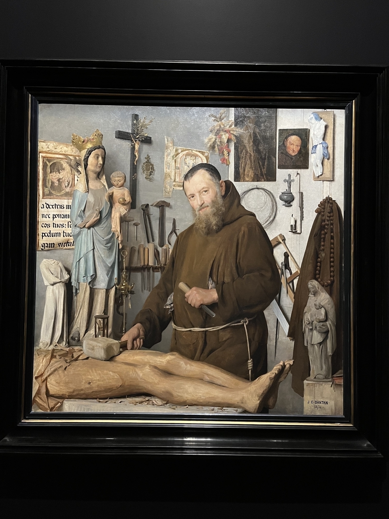 A bearded monk chisels a wooden figure in a workshop, surrounded by religious statues, tools, and crucifixes. Latin text partially reads: “a dextris… nec pomæ… pedum…” Artist signed: “J. E. Dantan, 1874.