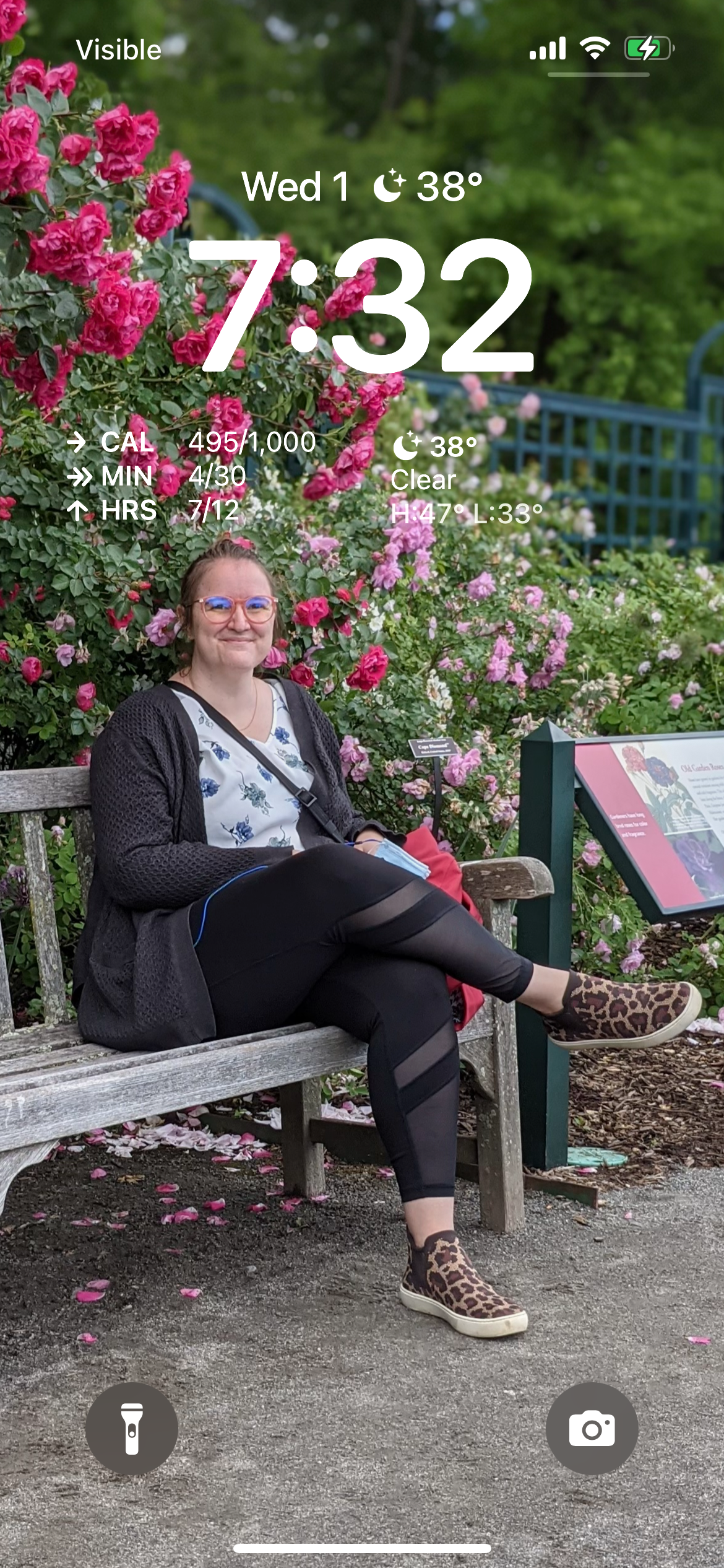 A person sits on a wooden bench, smiling, amidst vibrant pink roses. Surrounding them is a garden with informational signage. The screen displays the time as 7:32 and a temperature of 38° with other phone data.