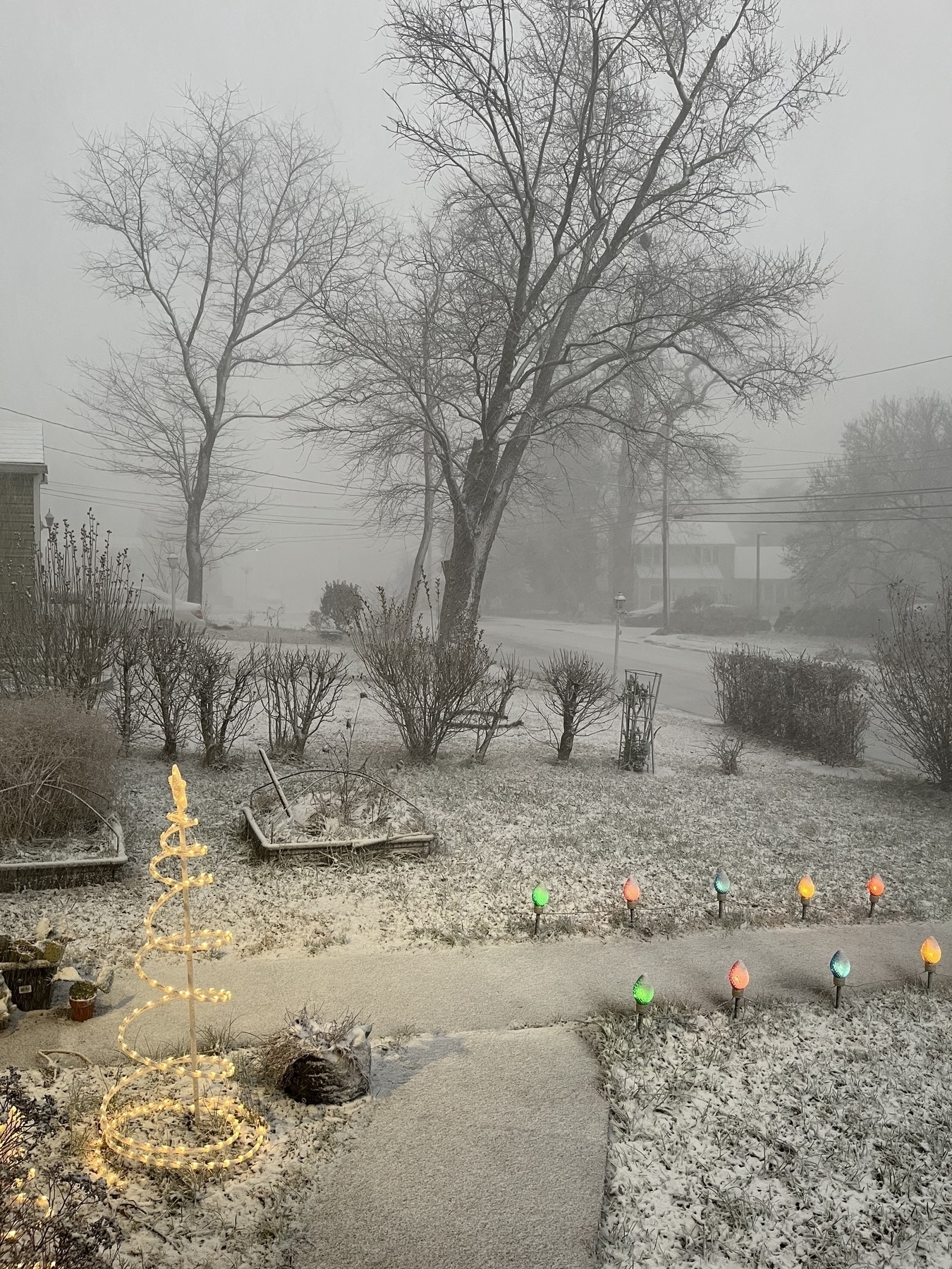 A spiral light structure glows warmly in a snowy front yard, surrounded by colorful path lights. Bare trees and bushes are covered in snow during an overcast winter day.