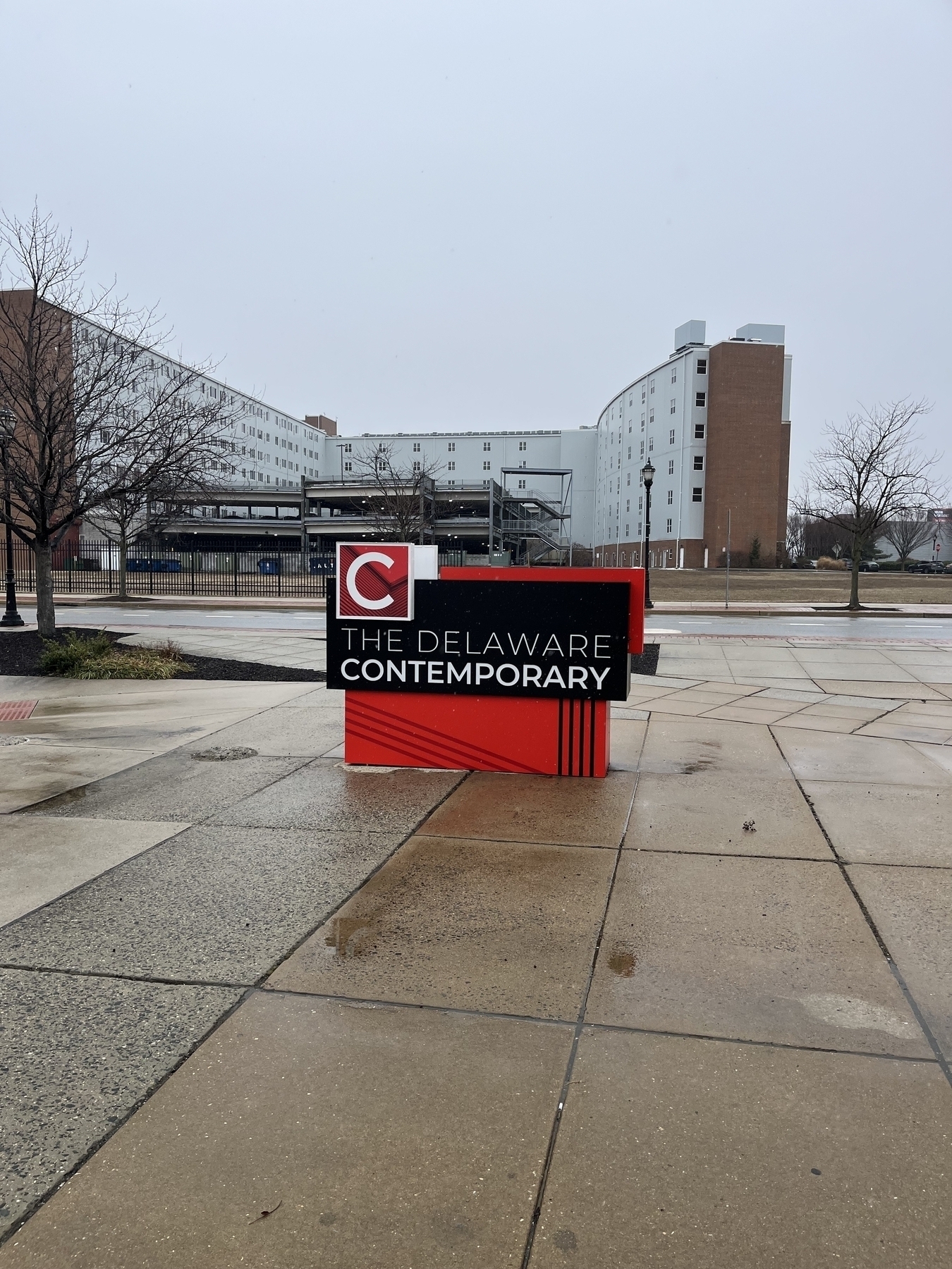 A red and black sign displaying “The Delaware Contemporary” stands on a wet pavement, surrounded by modern buildings and bare trees on a cloudy day.