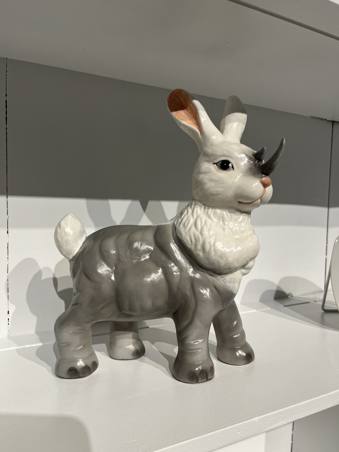 A ceramic figurine with a rabbit’s head and rhino body stands on a white shelf, blending animal features creatively. It has large ears and a prominent horn.