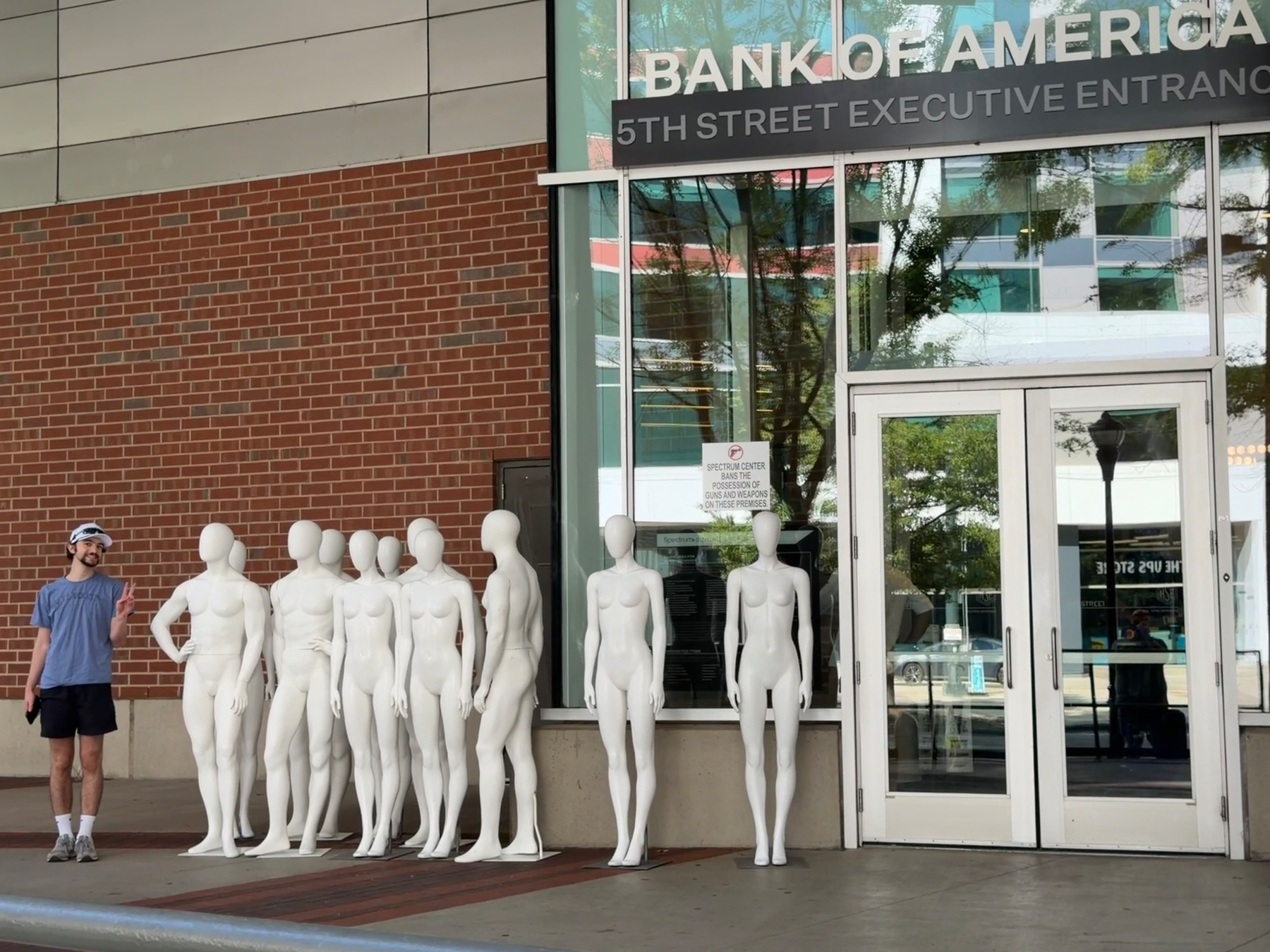 A group of white mannequins stands upright in front of a brick wall next to a smiling person. The entrance reads Bank of America 5th Street Executive Entrance of the Spectrum Center. 
