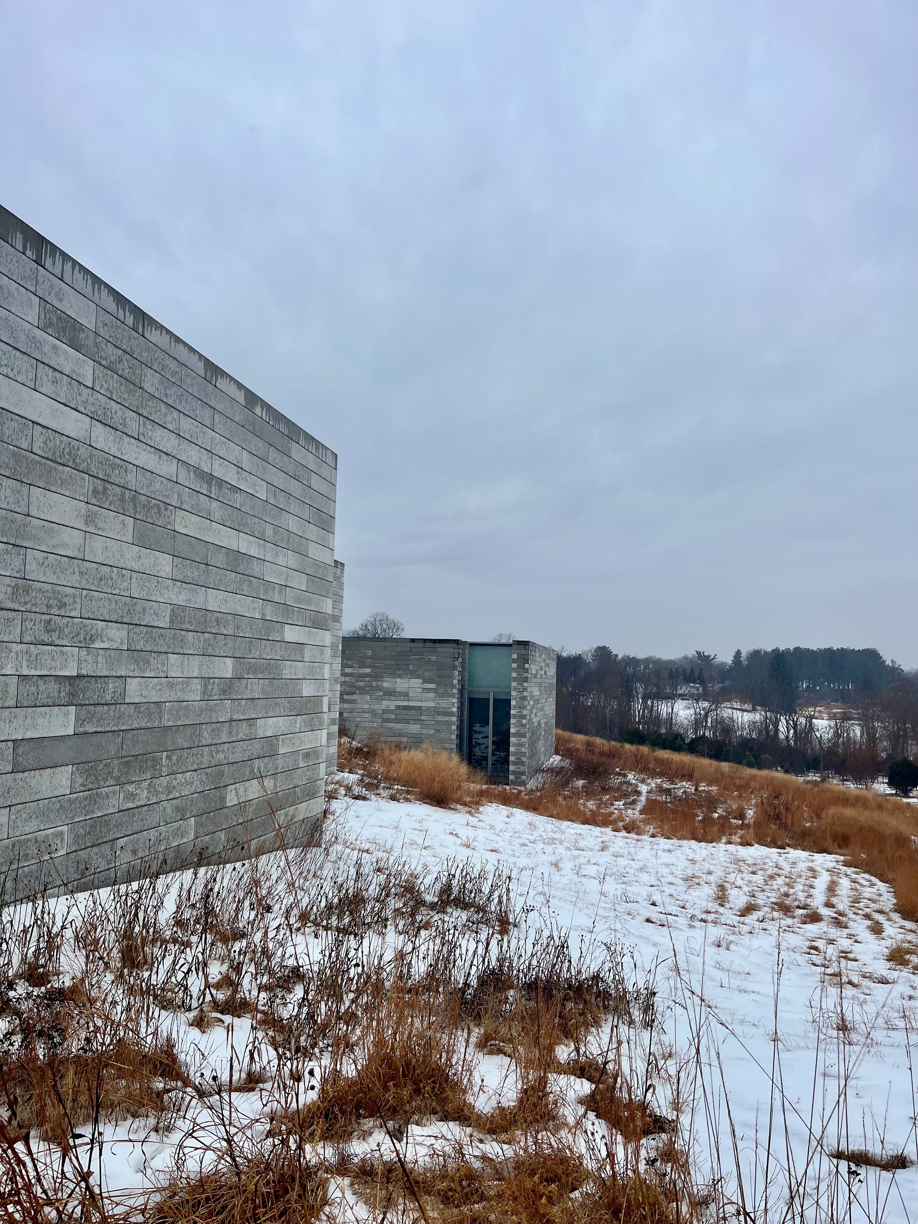 Stone buildings stand amidst a snowy, grassy field under a cloudy sky, with distant trees visible in the background.