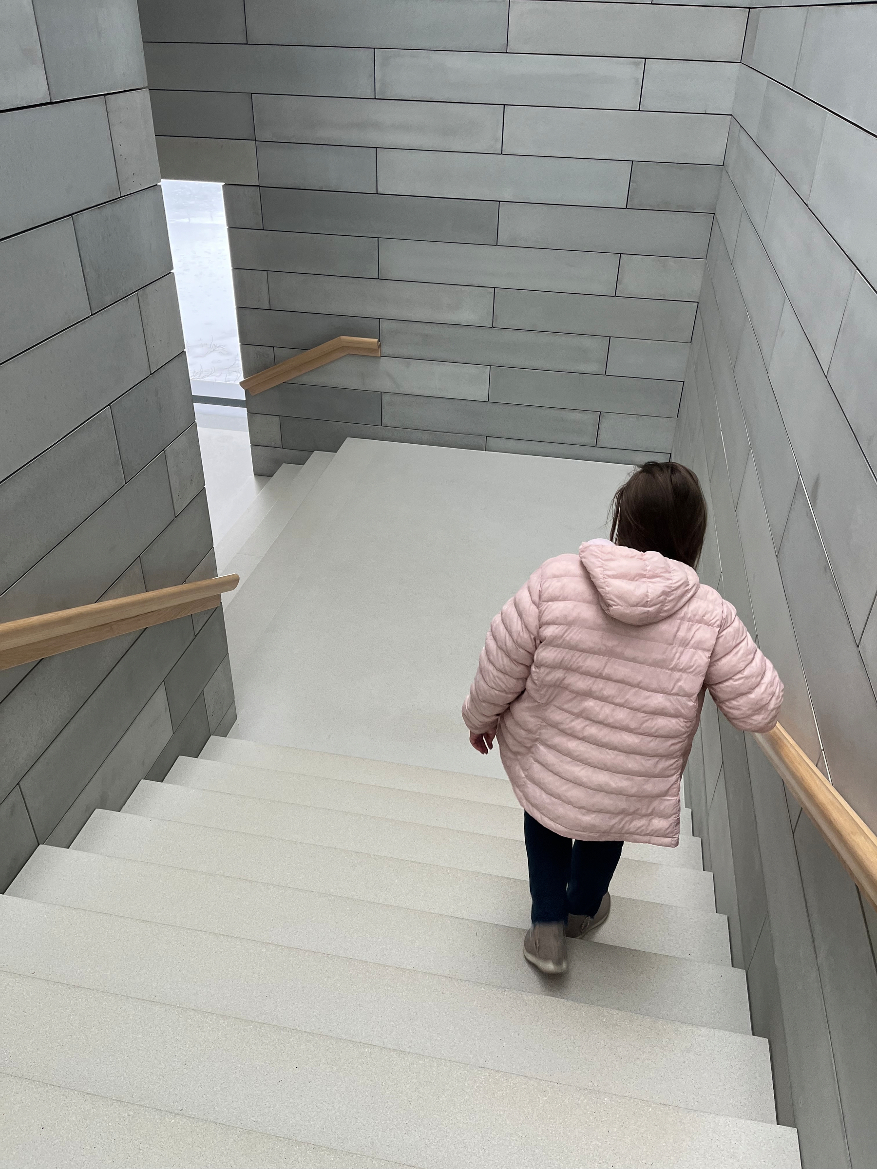 A person in a pink puffy jacket descends wide, light gray stairs bordered by light wooden handrails, within a minimalist gray-tiled stairwell. Bright natural light filters through a window.