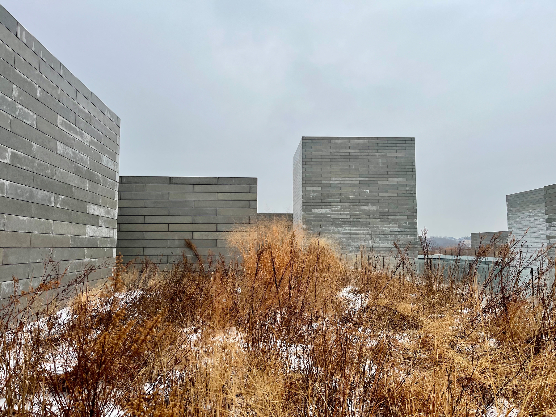 Concrete walls stand amidst dried grass, with patches of snow scattered. The setting appears overcast, creating a stark, minimalist landscape, emphasizing the geometric structure and natural elements.