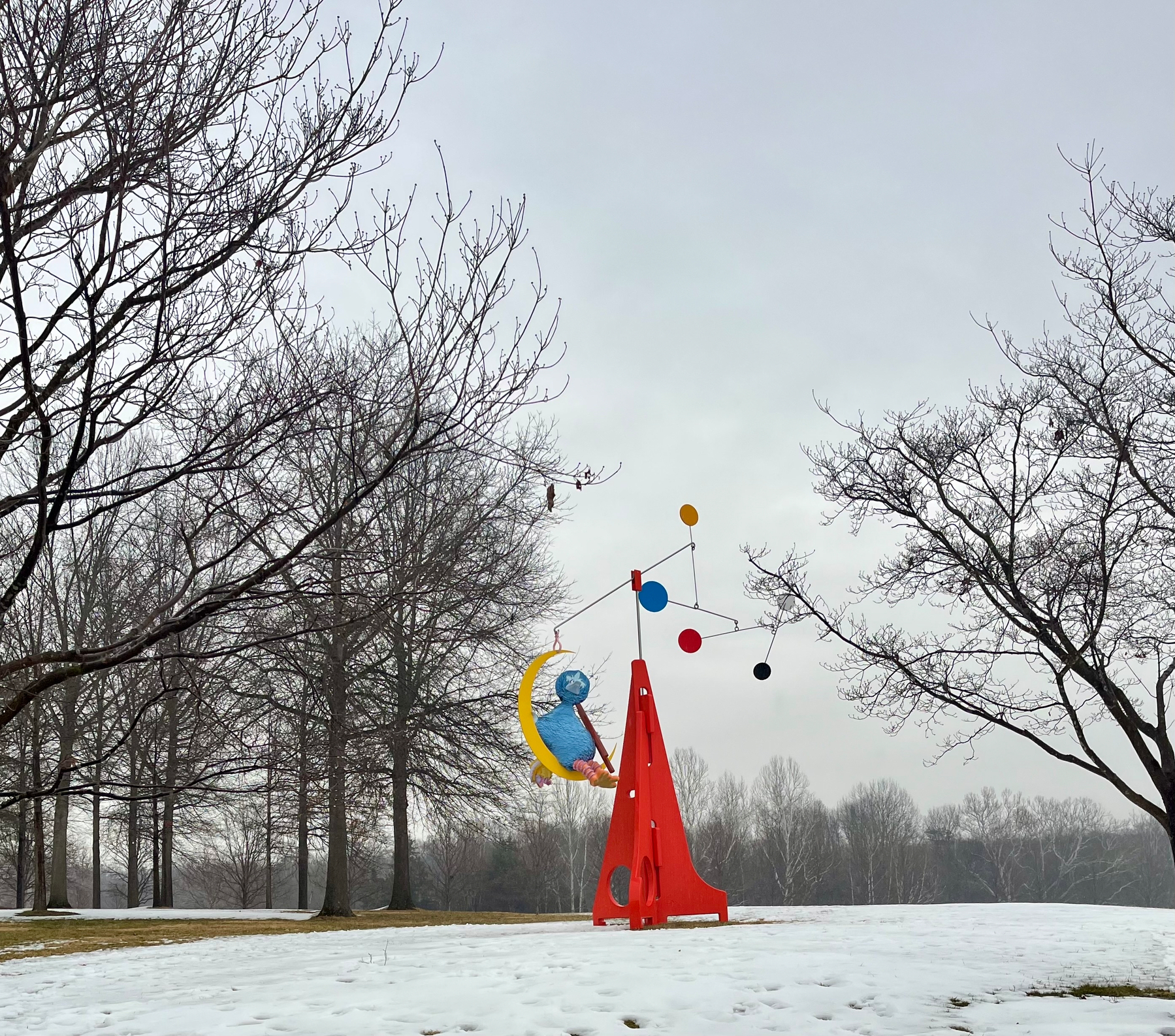 Colorful abstract sculpture with a red base balances various shapes, set on a snowy landscape surrounded by leafless trees under a cloudy sky.
