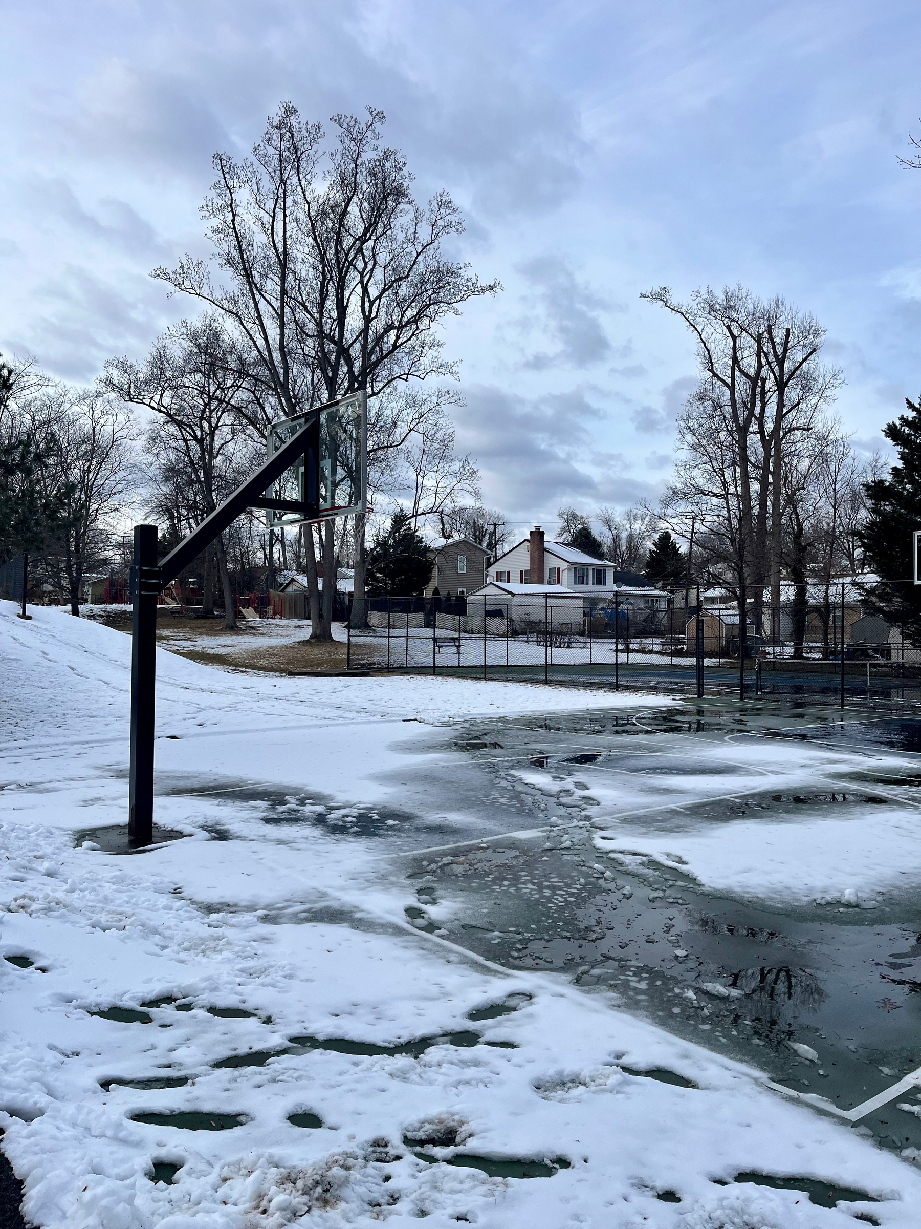 Basketball hoop stands idle on a snow-covered court with a layer of ice, surrounded by leafless trees and residential houses under a cloudy sky.