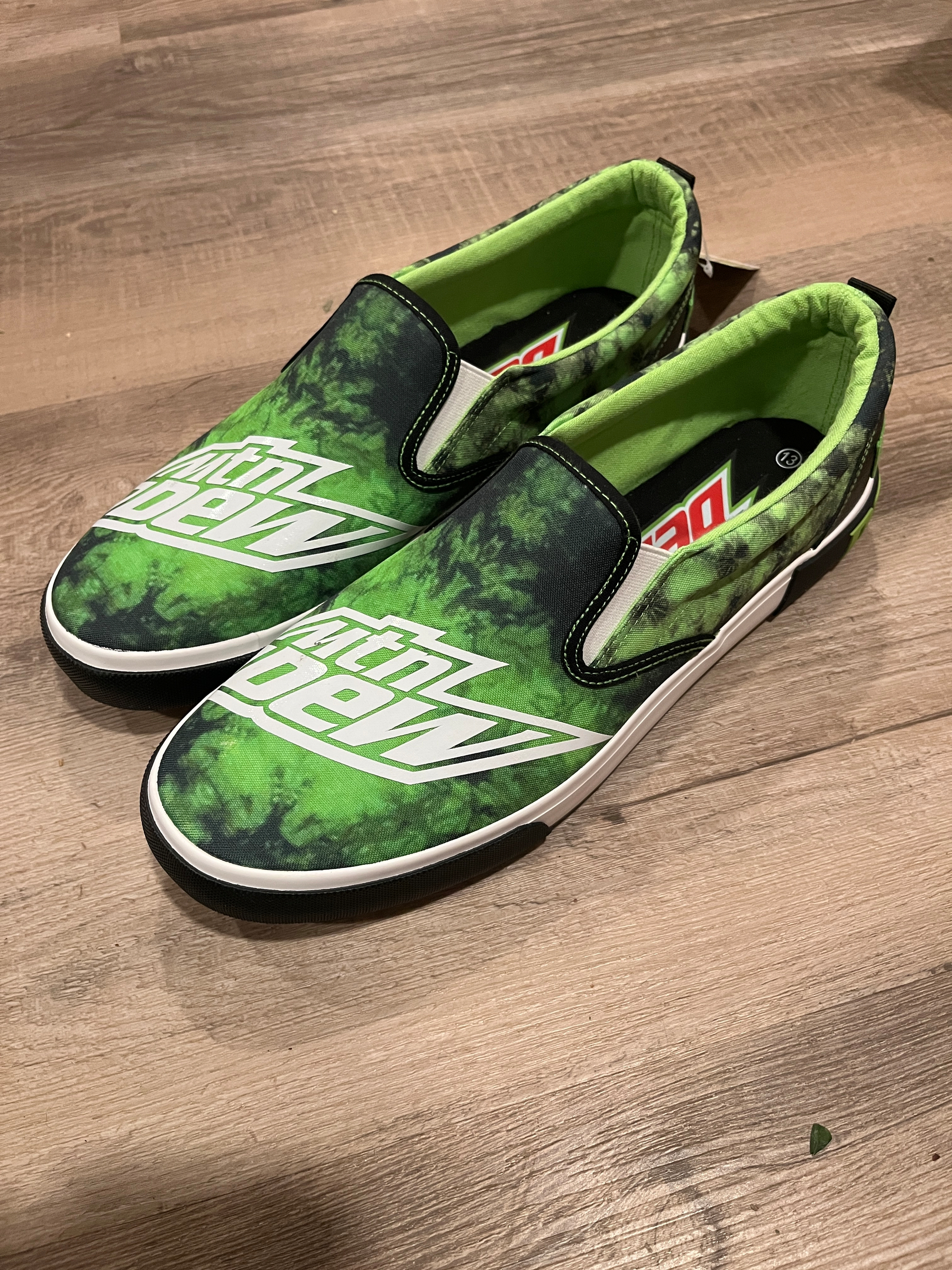 Green slip-on sneakers display the Mountain Dew logo, positioned on a wooden floor. The shoes feature a vibrant, tie-dye pattern with a soft green interior lining.