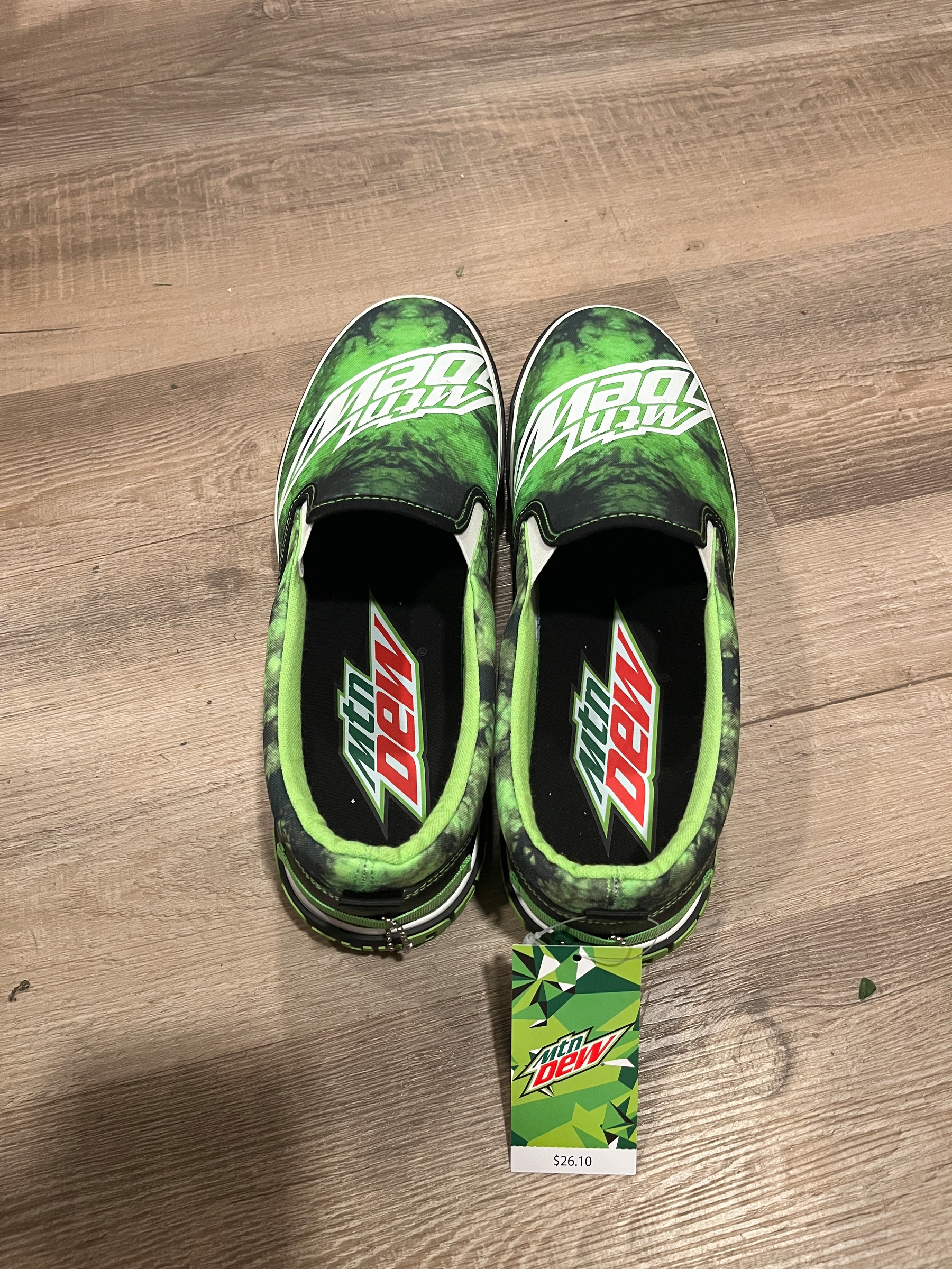 Green slip-on shoes display the Mountain Dew logo, resting on a wooden floor. A price tag attached reads: 26.10, themed similarly with the logo and colors.