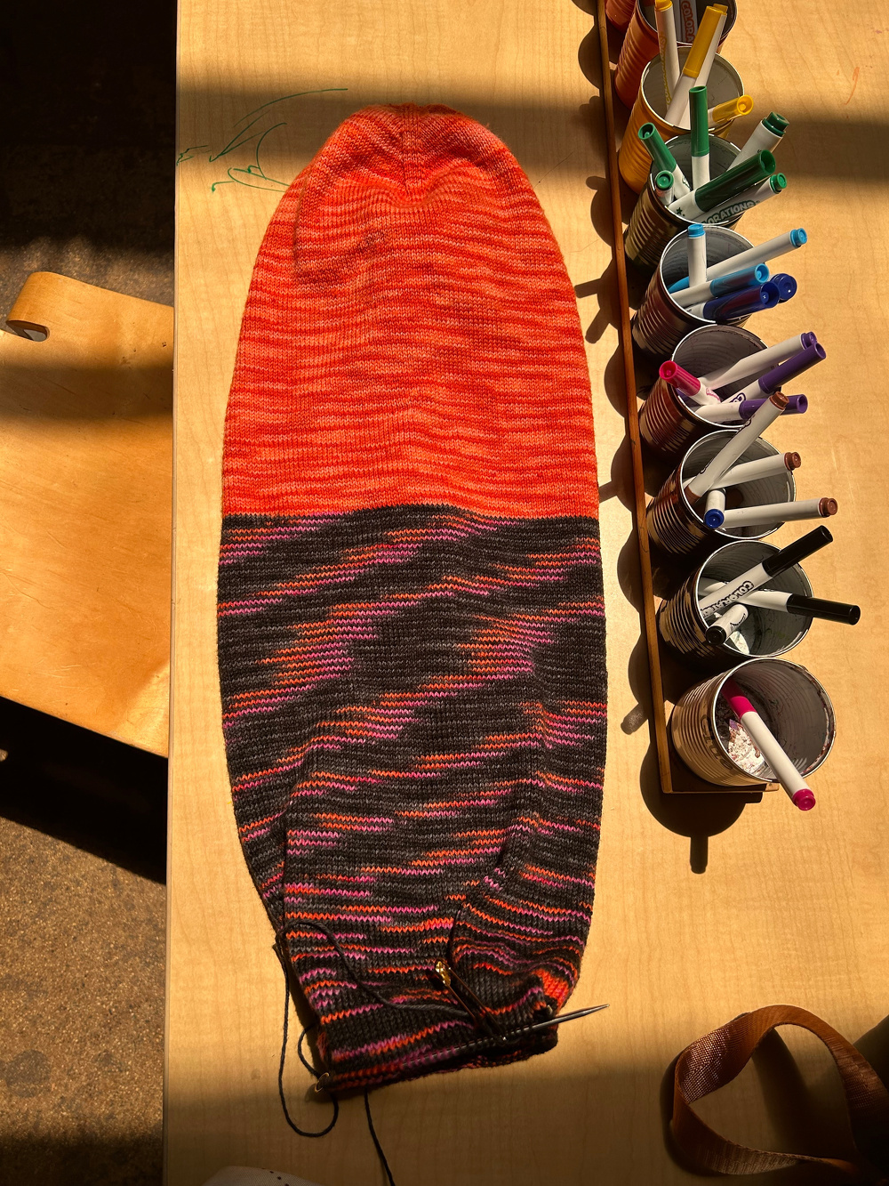 A large, colorful knitted piece with shades of orange and dark hues is laid on a table next to a row of cups holding various colored markers.