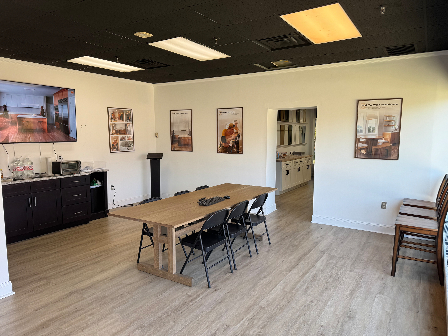 A modern office space features a wooden table with folding chairs, wall-mounted posters, and a large TV, complemented by light wood flooring and black ceiling accents.