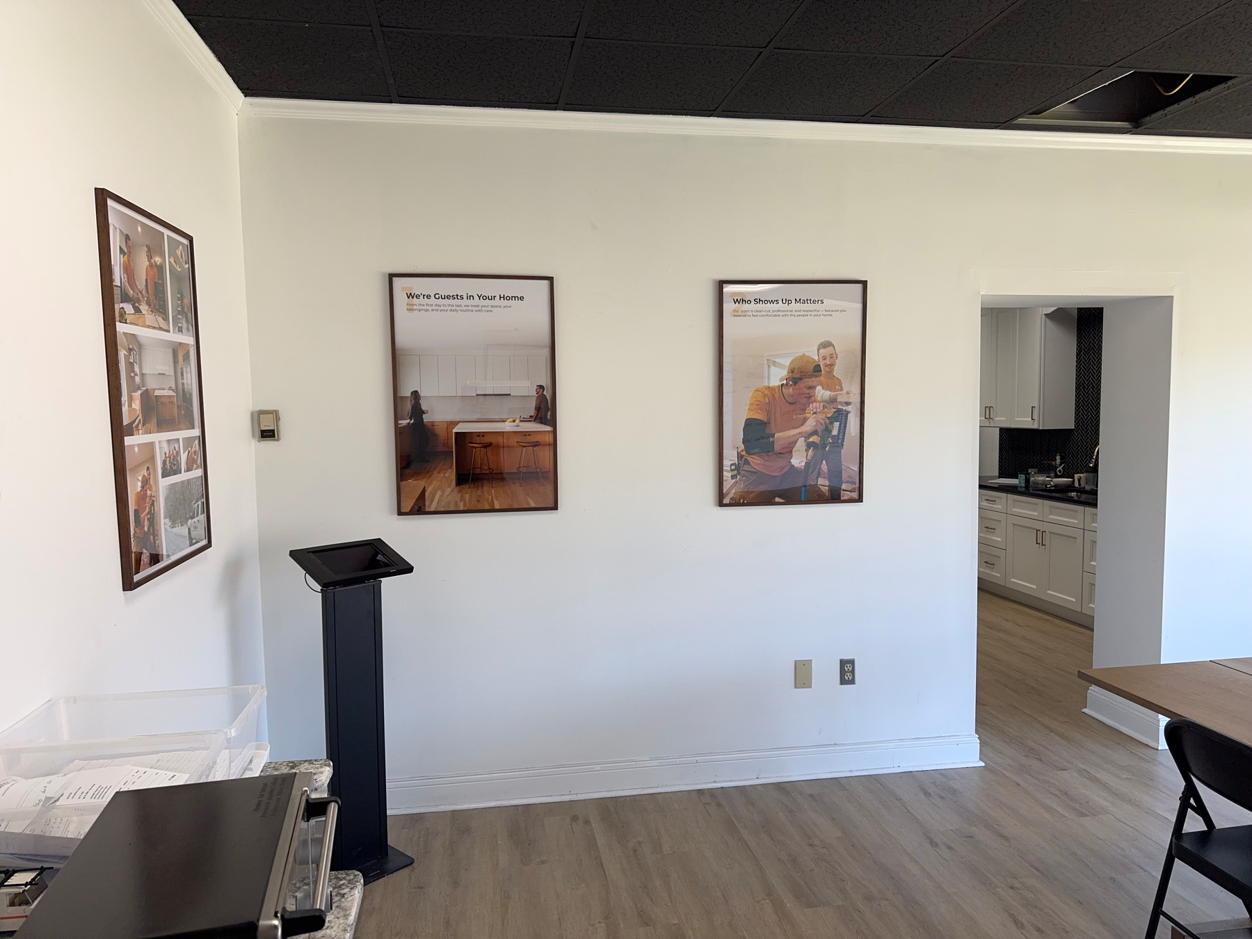 A room features a stand, framed posters on the wall, and an open doorway leading to a kitchen area.