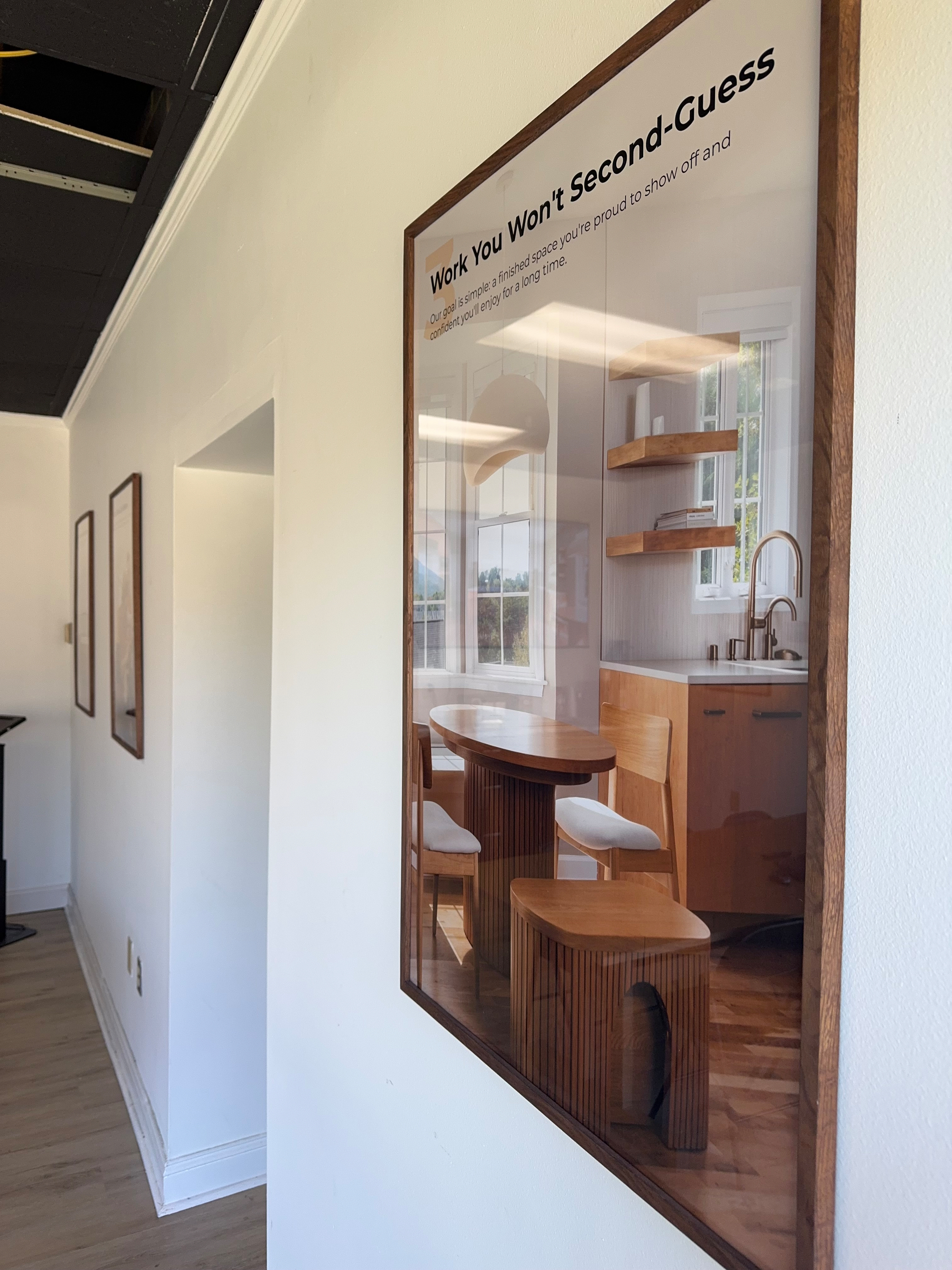 A framed poster showcasing a stylish kitchen interior with wooden furniture is mounted on a white wall in a corridor.