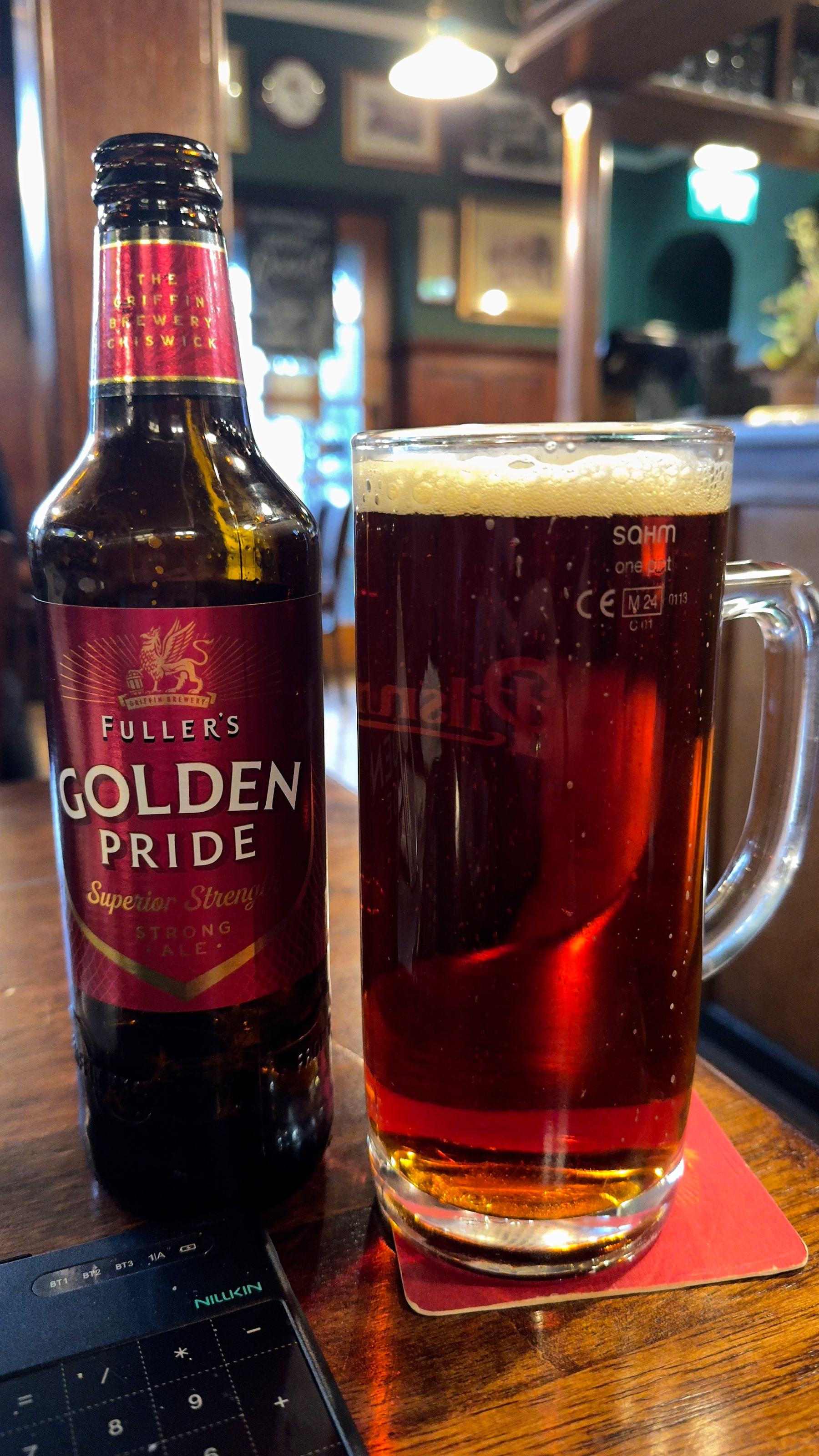 A bottle and mug of Fuller's Golden Pride beer sit on a wooden table in a cozy pub setting.