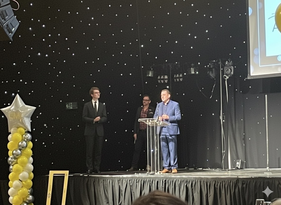 Three people in formal attire are on a stage with a starry backdrop, and one is speaking at a clear podium.