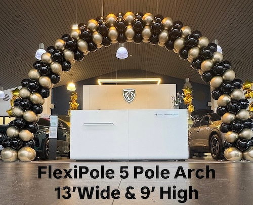 A balloon arch made of black and gold balloons is displayed in a car showroom, flanked by two parked cars.