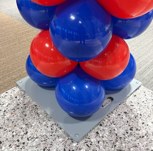 Shiny red and blue balloons are grouped together, anchored to a gray base on a speckled surface.