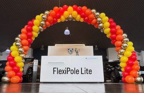 A vibrant balloon arch of red, orange, yellow, and gold frames a display for FlexiPole Lite at an indoor event.
