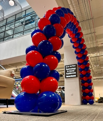 A colorful balloon arch made of alternating red and blue balloons is set up in a bright indoor space near a couch and a sign for technology and home appliances.