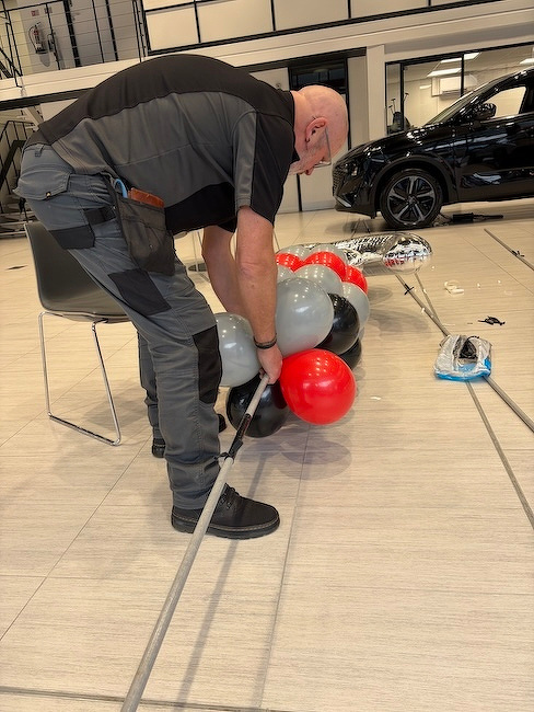 A person is arranging red, gray, and black balloons in a room with a car in the background.