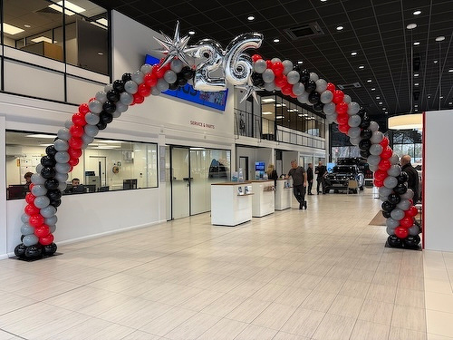 A balloon arch with black, red, and gray balloons, featuring the number 26, is set up in a modern showroom with people in the background.