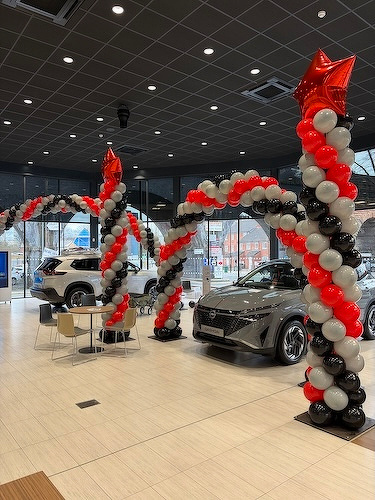 A car showroom is decorated with red, black, and white balloon arches, and features two parked cars and a small seating area.