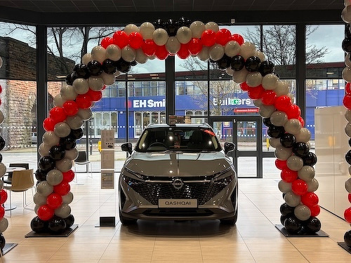 A car is displayed in a showroom, framed by an arch made of red, black, and gray balloons.