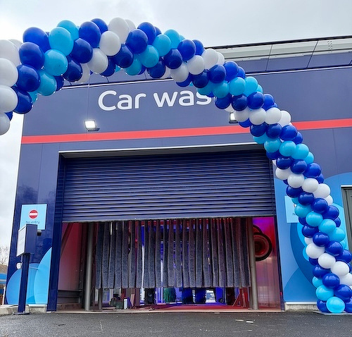 A car wash entrance decorated with a colorful arch of blue, white, and light blue balloons.