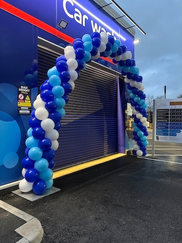 A colorful balloon arch in shades of blue and white is set up outside a car wash entrance.