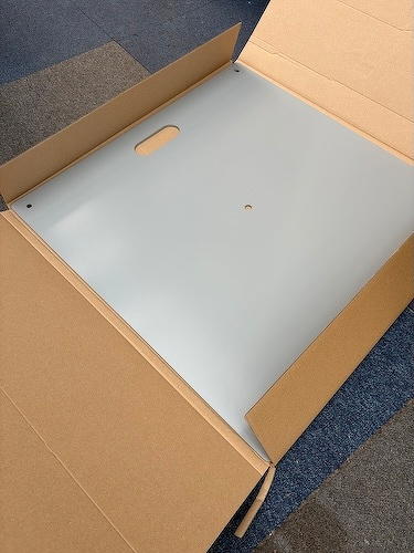 A cardboard box contains a flat, silver metallic panel with a handle and a small hole.