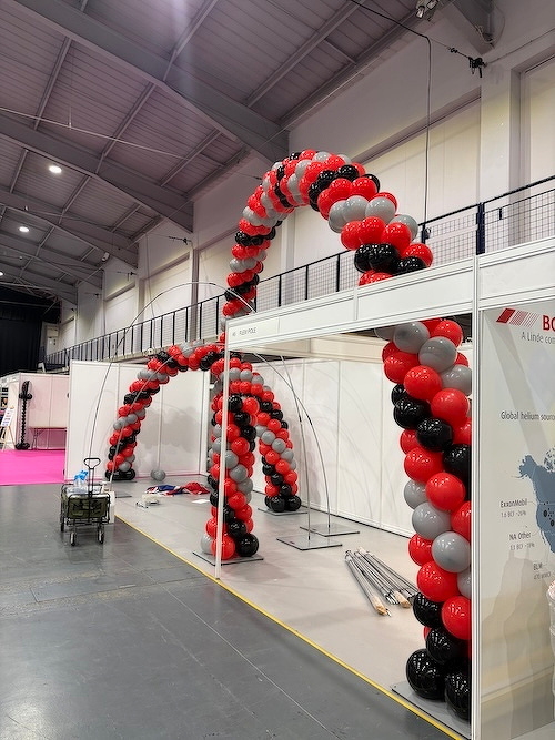 A large archway made of red, black, and gray balloons stands inside a spacious indoor venue.