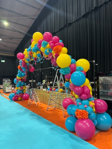 A vibrant arch made of yellow, pink, and blue balloons stands over a platform with ladders and equipment nearby.