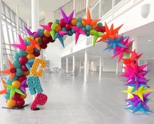 A colorful balloon arch with star-shaped decorations, letters GRA and a peace sign, stands in a spacious, modern indoor setting.