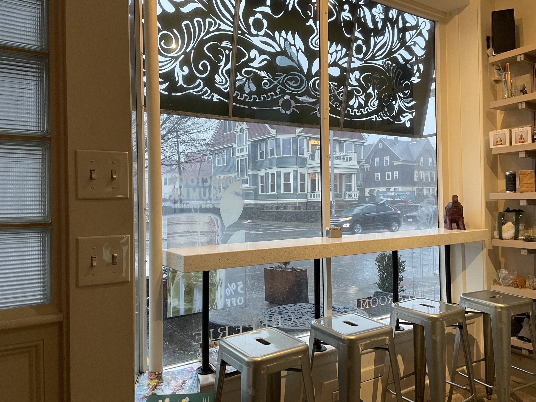 Looking outside from in, a row of stools beneath a shelf at a window with a metal lace awning. Outside, the street, car and houses are being drenched in driven rain. 