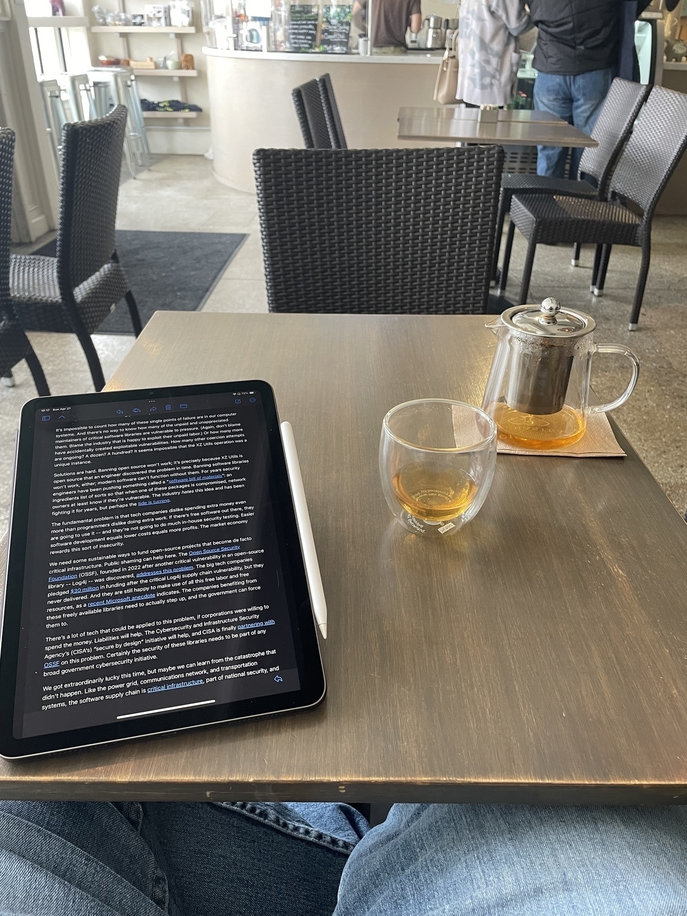 An iPad with text on the screen is angled towards the viewer on the left of a table. On the right, a nearly empty clear mug and tea sit. In the background, an empty chair faces the viewer on the opposite side of the table, and beyond that, more tables, chairs, and a cafe counter with people ordering. 