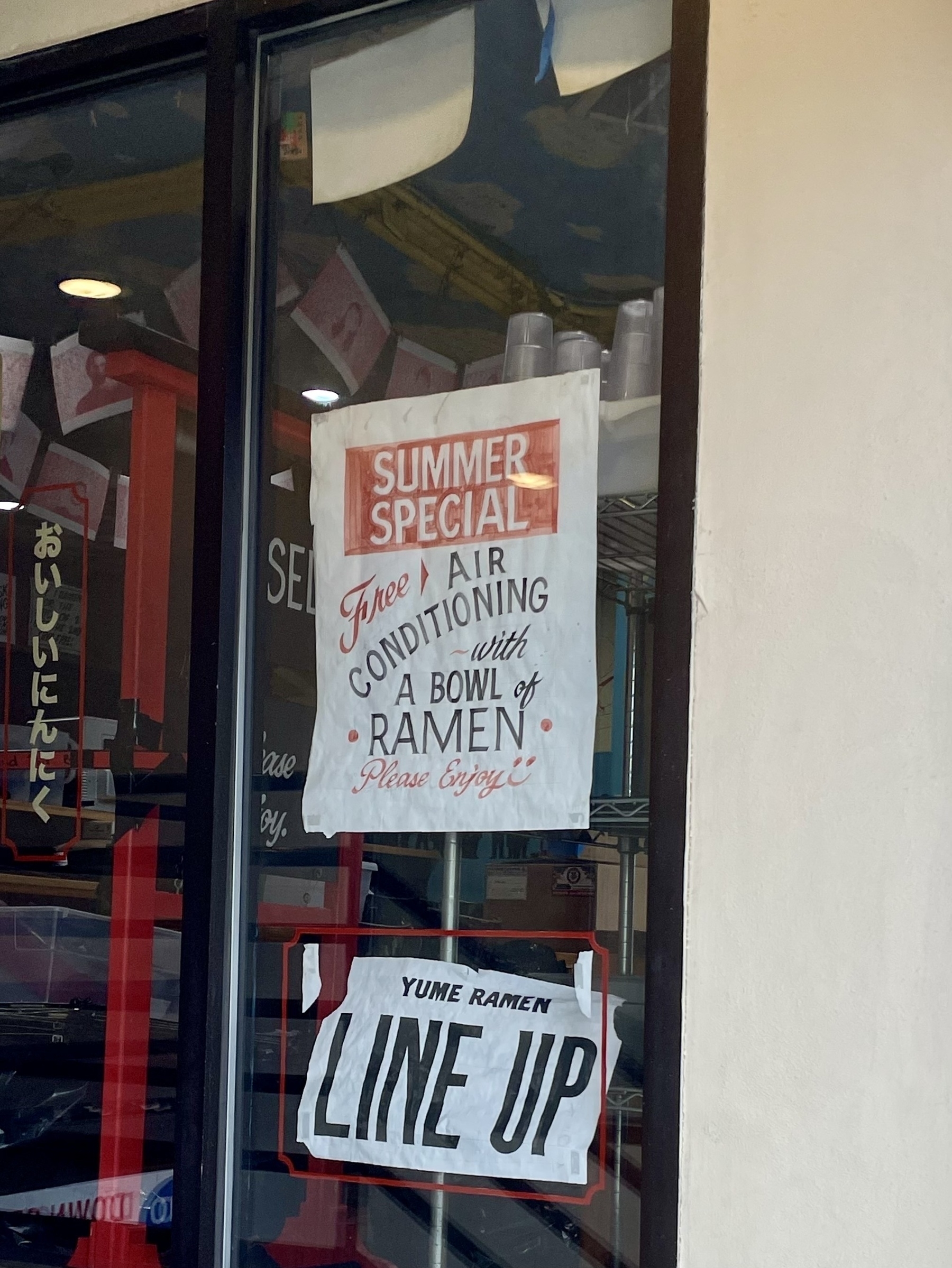 A sign in the window of a ramen shop advertises in stylized script, 'Summer Special: Free Air Conditioning with a bowl of raman. Please enjoy!'