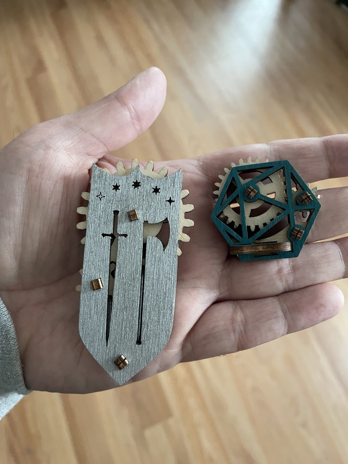 On a palm facing the camera, two flat wooden fidget spinners, one grey in the shape of a shield with axe and sword cutouts, the other a green hexagon with lines implying a die. Each have gears sticking out of edges that can be spun by fingers. 