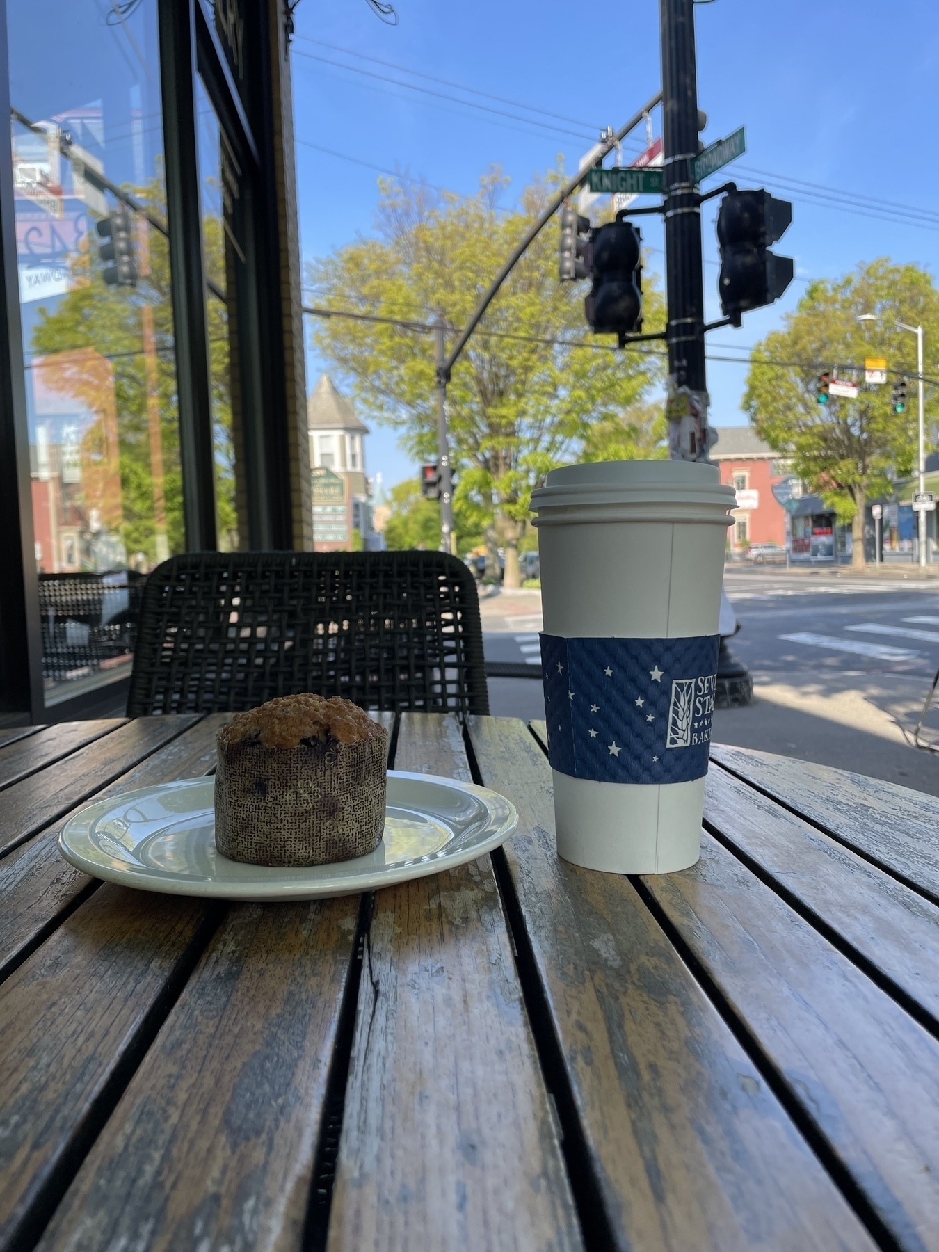 A berry muffin and a large paper to-go cup sit in a cafe table outside, in the shade. Behind it is another table, a street light pole, trees, houses and sky. 