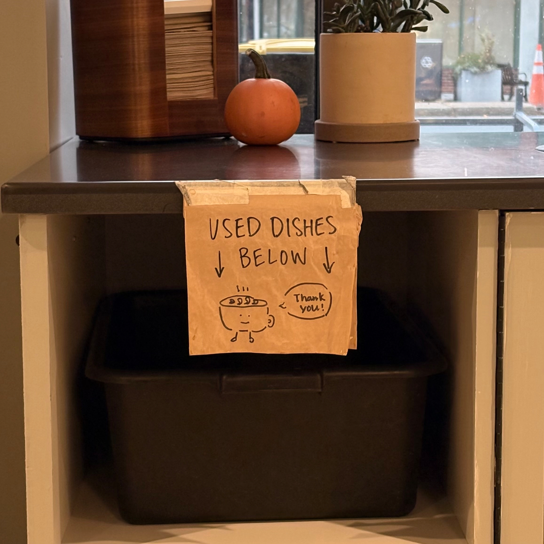 A counter with a napkin holder, a small pumpkin, a potted plant, and a sign indicating a bin below for used dishes. The sign contains a drawing of a coffee mug with legs saying, “Thank you!”