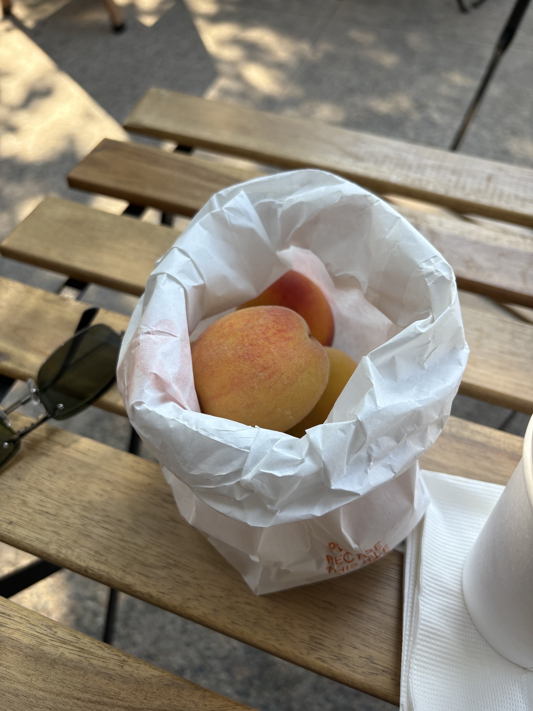 On a shaded, slatted wood table, an open white bag holds some small, fresh peaches. A pair of sunglasses lays folded behind the bag. 