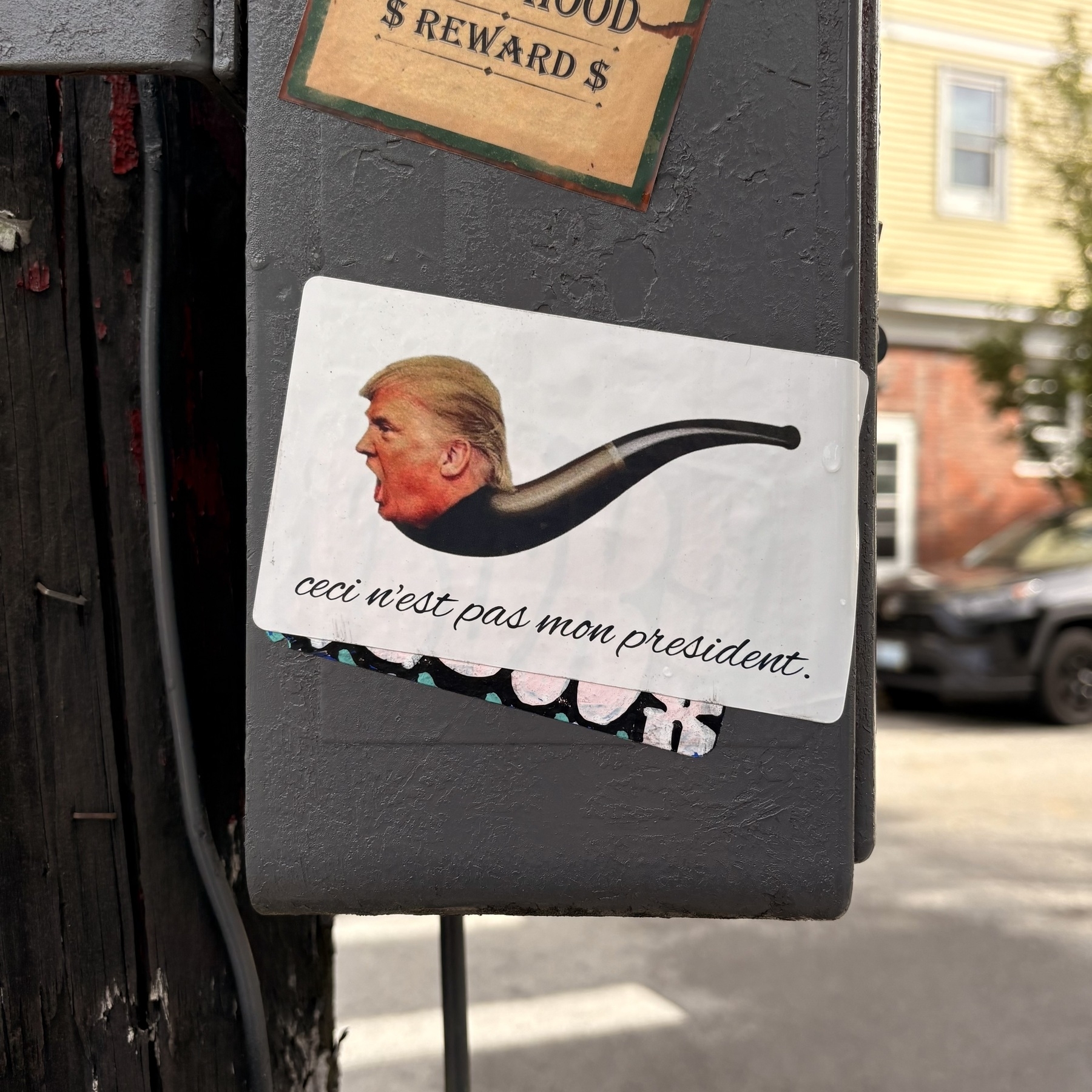 On a white sticker on a grey utility pole box, is a parody of a famous painting: Trump’s head at the end of an old fashioned pipe, with cursive text below it reading, “ceci n’est pas mon president.”