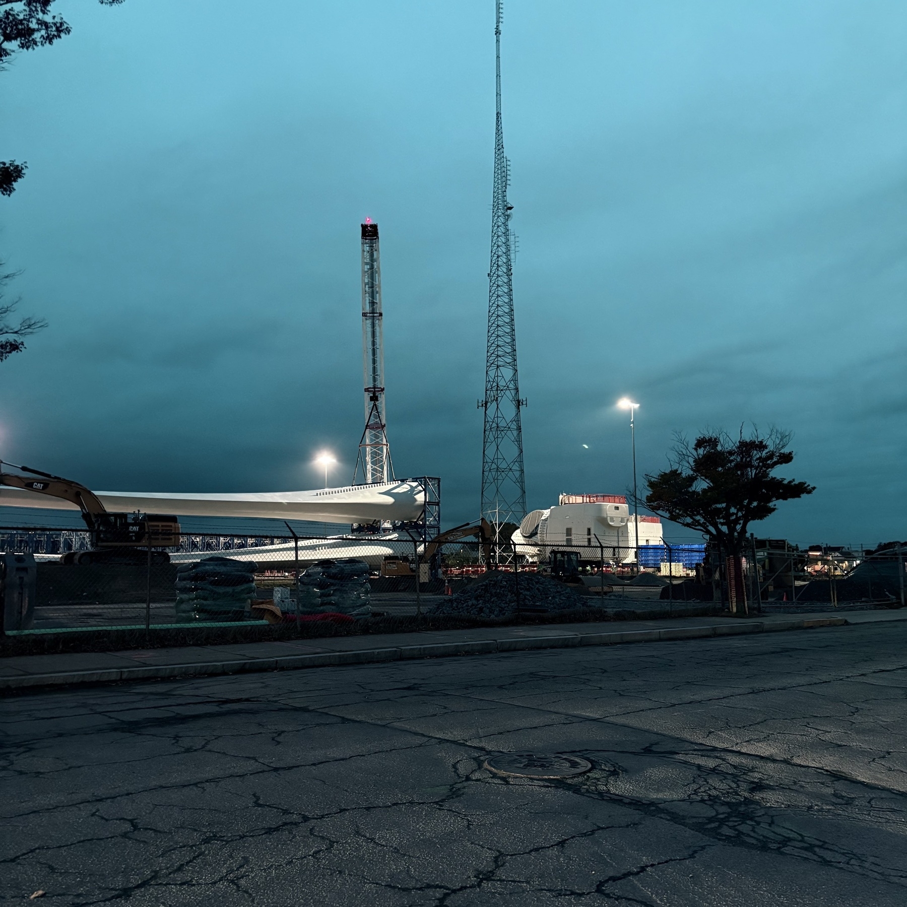 Across a street and behind a chain link fence, two white industrial windmill blades lay held flat in construction pipes. In front are some construction vehicles, behind are two radio towers and a white warehouse building, all under an overcast twilight sky. 
