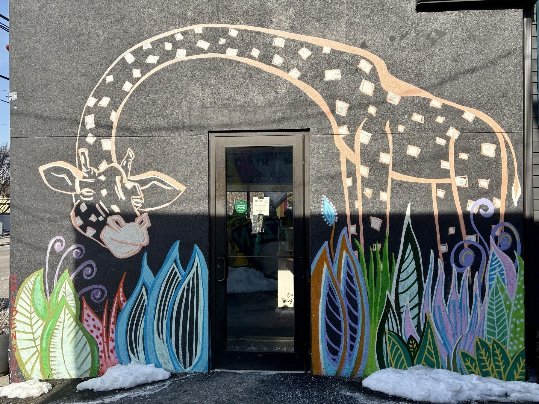 A colorful mural of a giraffe surrounded by tall, leafy multi-colored plants decorates the black wall around a door.​